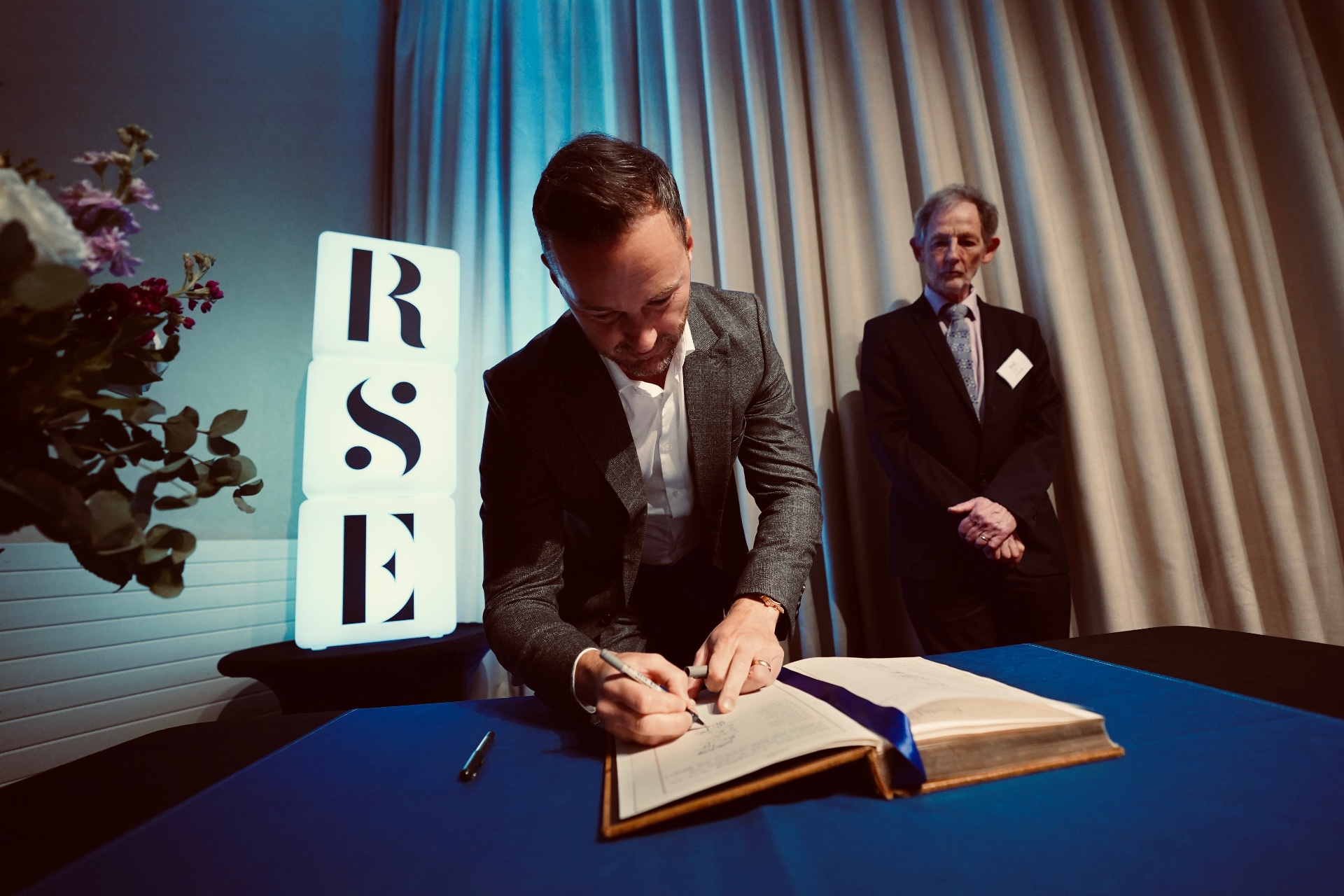 Michael Welch signing the Royal Society of Edinburgh roll of Fellows at an admission ceremony, with the RSE logo lit behind him.