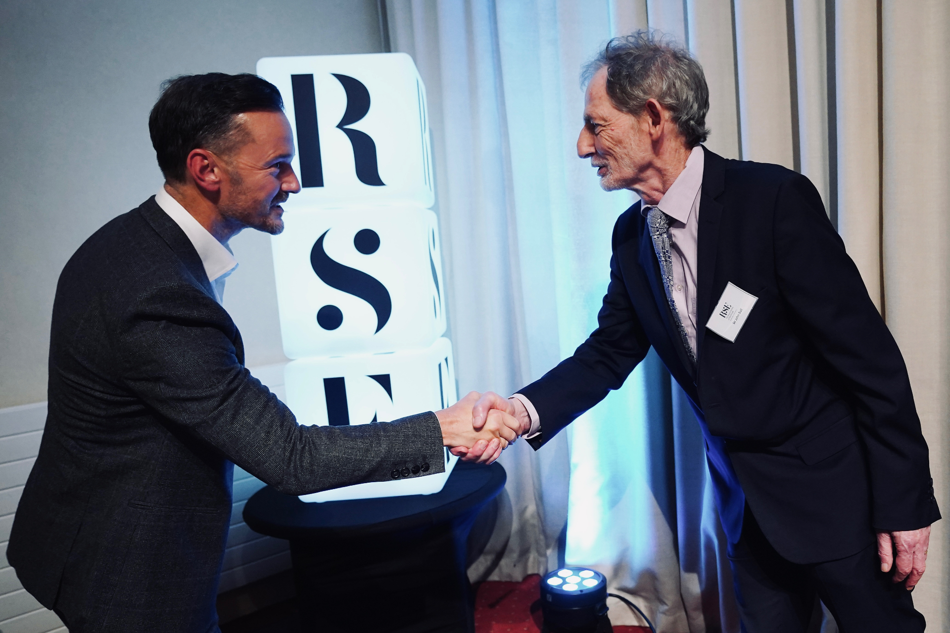 Michael Welch shaking hands with a Royal Society of Edinburgh officer beside the illuminated RSE logo.