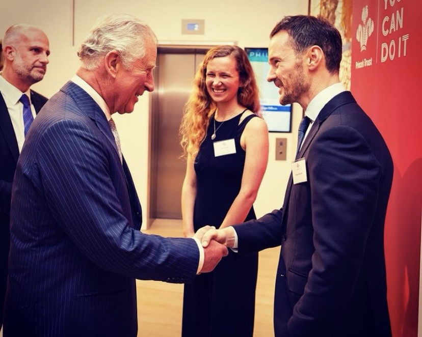 Michael Welch shaking hands with the then Prince of Wales at a Prince's Trust event, beside a 'You Can Do It' Prince's Trust banner.