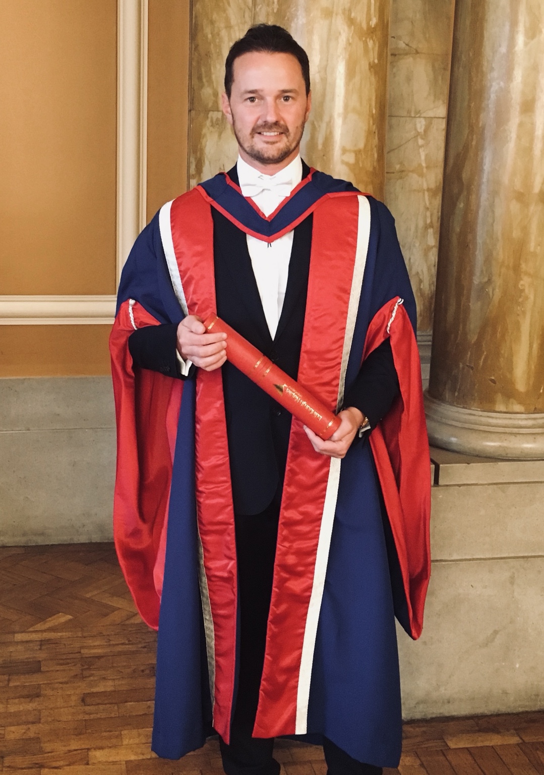 Michael Welch in navy and red academic robes, holding the scroll for his honorary Doctorate of Enterprise.
