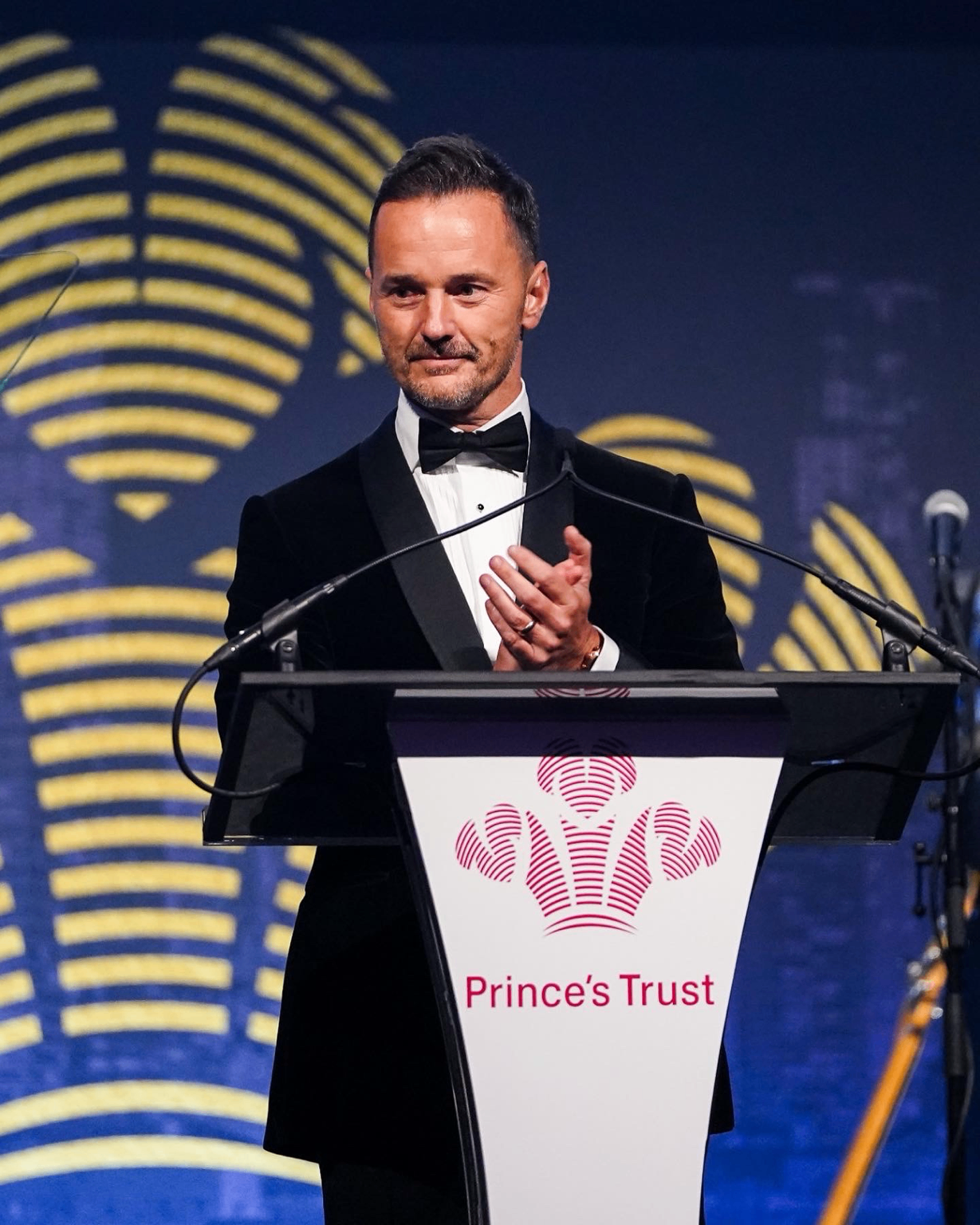 Michael Welch in black tie, applauding at a lectern bearing The Prince's Trust logo.